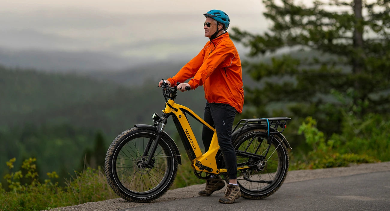 A person wearing an orange jacket and a blue helmet riding a yellow electric bicycle on a path with a scenic view of a forested area in the background