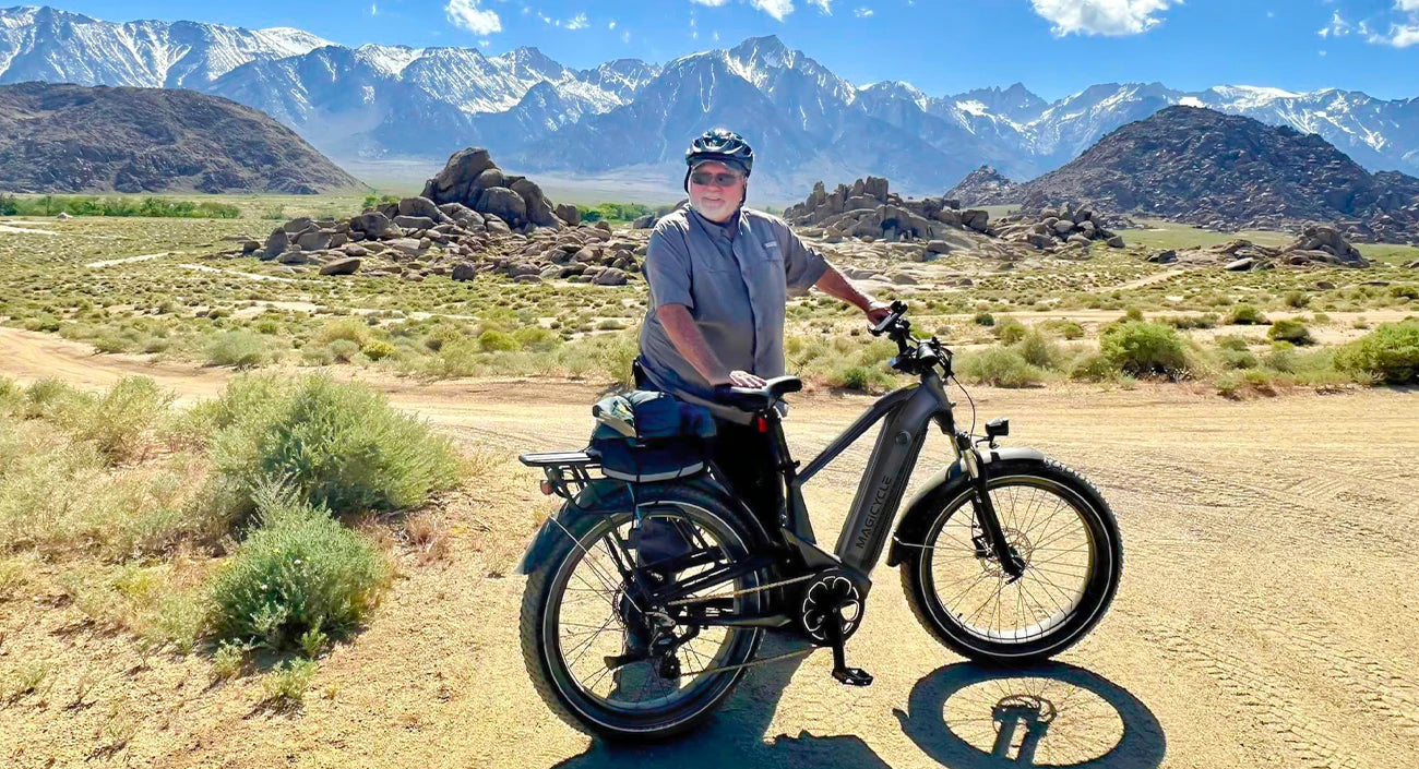 An elderly man stands behind an electric bicycle with mountains and hills and low bushes behind him