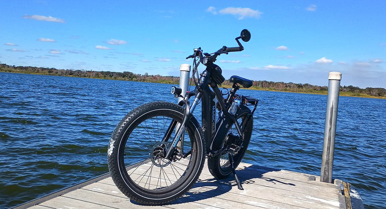 An electric bicycle parked on a wooden dock overlooking a body of water with trees and a blue sky with scattered clouds in the background